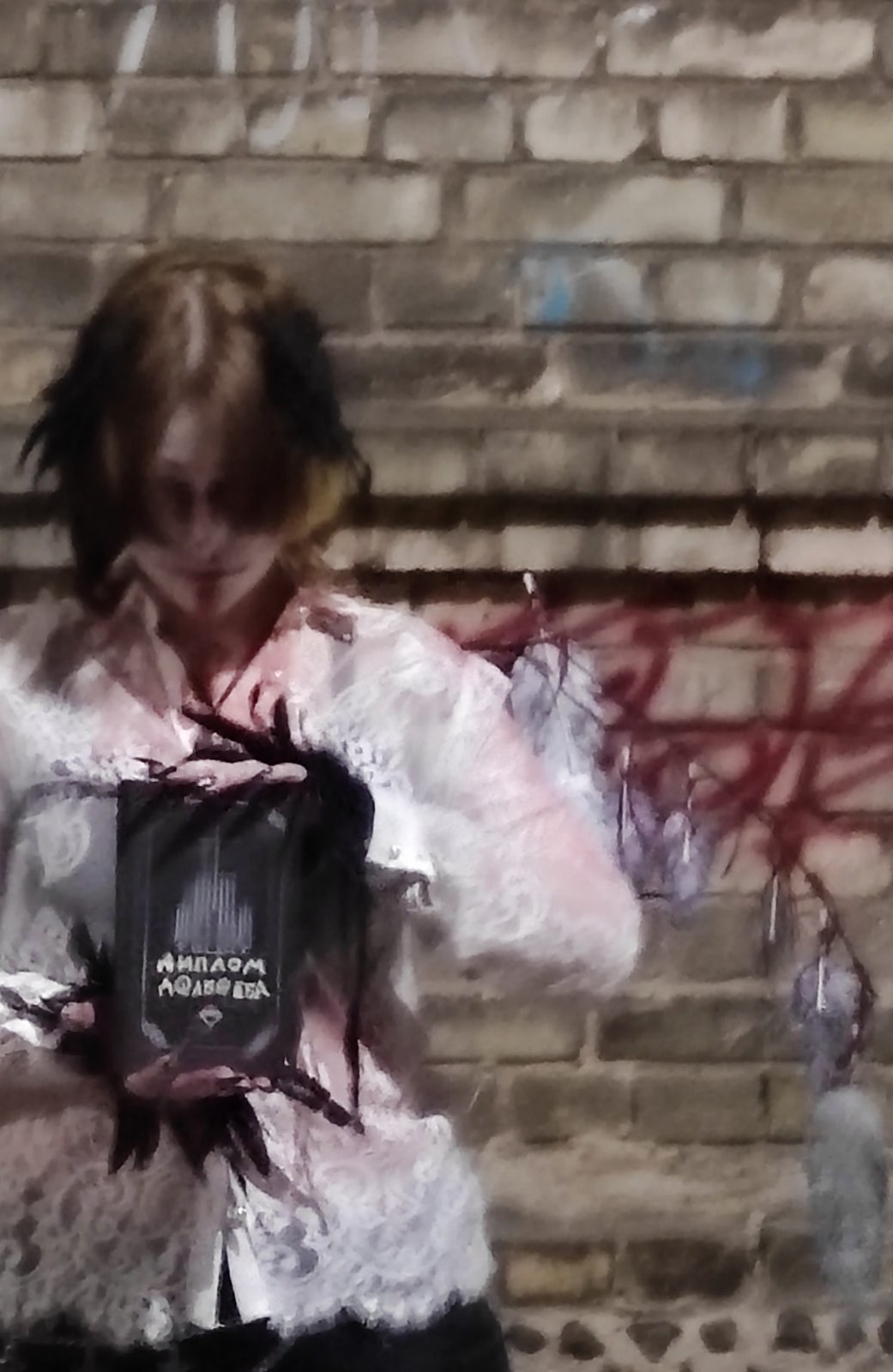 Figure in white lace with feathers holding a handwritten expulsion order against a brick wall